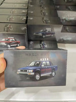 A hand holds a miniature model car box featuring a blue Mazda pickup truck, with multiple identical boxes stacked in the background.