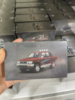 A hand holds a small rectangular product box featuring a red and black Masdi pickup truck illustration on a dark gray background. Multiple similar boxes are stacked in the background on a shelf.