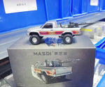 A silver toy model truck with orange and red decals and a mounted gun, displayed on top of its gray packaging box. The box features the text 'MAZDI' and 'TOYOTA' along with an illustration of the truck.