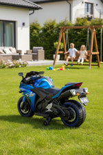 Blue electric toy motorcycle for kids on a green lawn with children playing in the background.