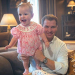 Man holding a baby wearing a 'Daddy's Girl' dress in a cozy living room.