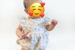 Baby wearing a white dress with ruffled shoulders, standing indoors.